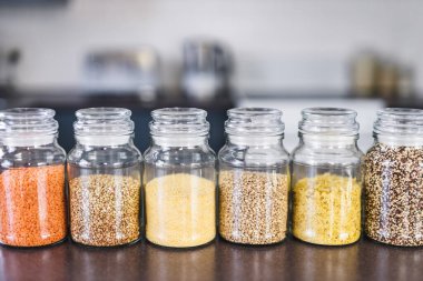 İçinde kinoa mercimek, karabuğday ve arpanın da bulunduğu farklı türde tahıl ve baklagillerin bulunduğu açık kiler kavanozları mutfak tezgahı, basit malzeme konsepti.
