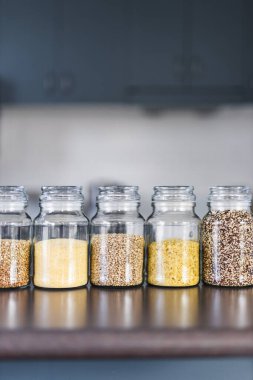 İçinde kinoa mercimek, karabuğday ve arpanın da bulunduğu farklı türde tahıl ve baklagillerin bulunduğu açık kiler kavanozları mutfak tezgahı, basit malzeme konsepti.