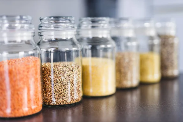 İçinde kinoa mercimek, karabuğday ve arpanın da bulunduğu farklı türde tahıl ve baklagillerin bulunduğu açık kiler kavanozları mutfak tezgahı, basit malzeme konsepti.