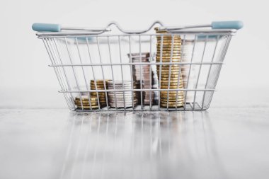 Alışveriş sepetinin içinde minimalist kompozisyonu, harcamaları ve tüketici davranışları ile sığ alan derinliğindeki beyaz arka planda çekilen farklı madeni paralar yığını 