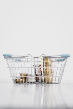 Alışveriş sepetinin içinde minimalist kompozisyonu, harcamaları ve tüketici davranışları ile sığ alan derinliğindeki beyaz arka planda çekilen farklı madeni paralar yığını 