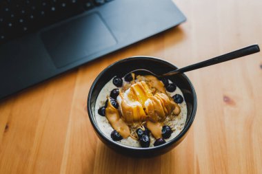 Evde dizüstü bilgisayarla çalışırken mango yulaf ezmesi yemek, uzaktan kumanda konsepti ya da ofiste öğle yemeği yemek.