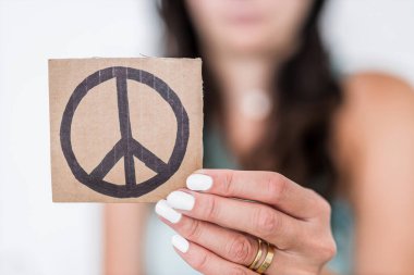Bir mukavva parçasının üzerinde barış işareti tutan kadın kamerayı sığ alan derinliğine, dünya barışı kavramına ve savaşların sona ermesine doğru çeviriyor.