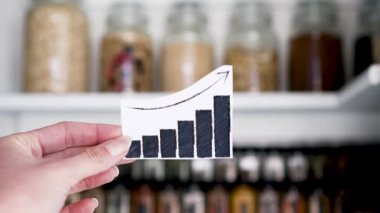 graphs showing prices of groceries going up in front of tidy organised pantry with jars and ingredients shot at shallow depth of field