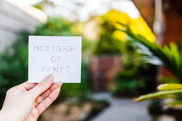 hand holding mortgage or rent sign in front of backyard and home exterior bokeh, concept of real estate affordability and property market