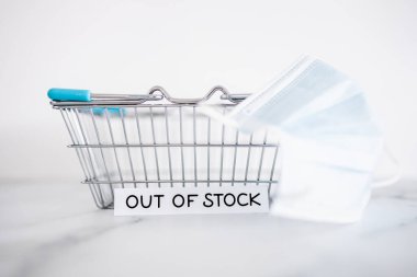 out of stock text in front of empty shopping basket with surgical mask on white background, concept of supply chain shortages and delays after the covid-19 global pandemic