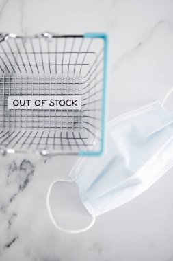 out of stock text in front of empty shopping basket with surgical mask on white background, concept of supply chain shortages and delays after the covid-19 global pandemic