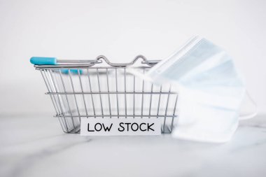 Low stock text in front of empty shopping basket with surgical mask on white background, concept of supply chain shortages and delays after the covid-19 global pandemic