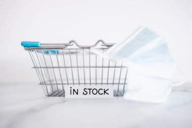 In stock text in front of empty shopping basket on white background with surgical mask, concept of supply chain shortages and delays after the covid-19 global pandemic