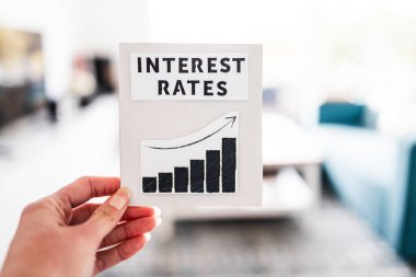 property affordabilikty and home loan payment increases, hand holding interest rates sign with graph showing stats going up in front of living room bokeh