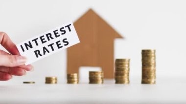 Interest Rates text with growing stacks of coins representing increasingly expensive loans and mortgages with cardboard house in the background, concept of global economy after the pandemic