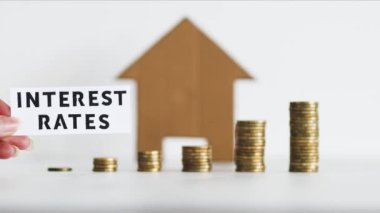 Interest Rates text with growing stacks of coins representing increasingly expensive loans and mortgages with cardboard house in the background, concept of global economy after the pandemic