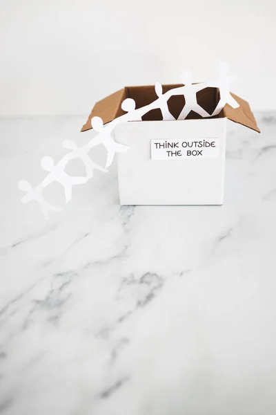 Think outside the box concept with paper people chain getting out of a white box with text on white marble	with minimalistic composition