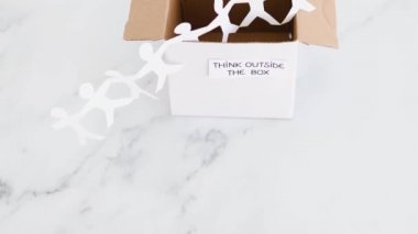 Think outside the box concept with paper people chain getting out of a white box with text on white marblewith minimalistic composition