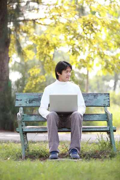 Молодой красивый мужчина с ноутбуком сидит на скамейке и смотрит в — стоковое фото