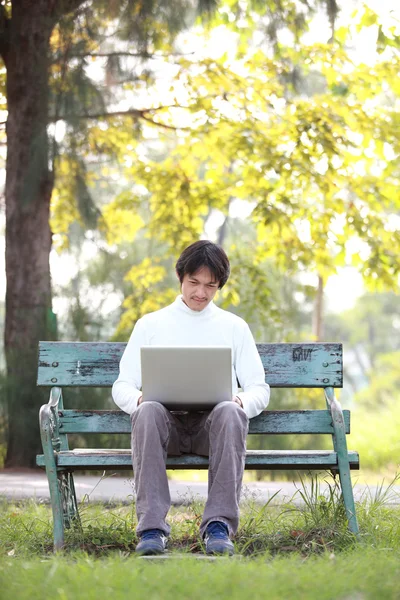 Un jeune homme beau utilisant un ordinateur portable assis sur un banc dans un parc . — Photo