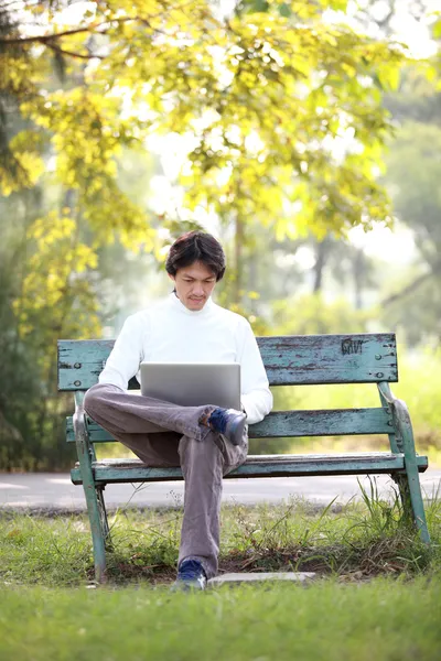 Młody, przystojny mężczyzna, za pomocą laptopa siedząc na ławce w parku. — Zdjęcie stockowe
