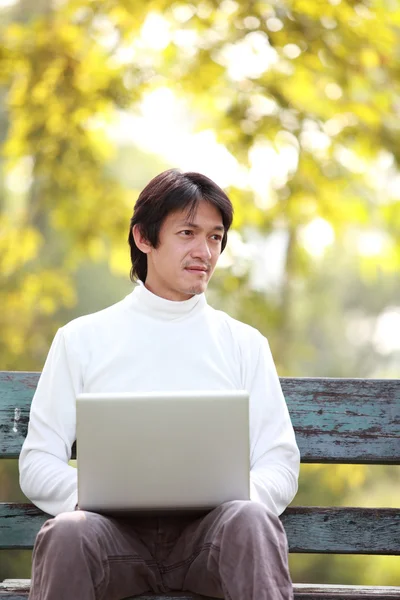 A young handsome man using laptop sitting on a bench looking in — Stock Photo, Image
