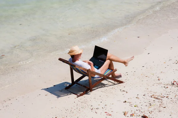 Mann sitzt mit Laptop auf einem Liegestuhl am Strand lizenzfreie Stockfotos