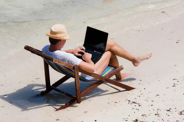 Mann sitzt mit Laptop auf einem Liegestuhl am Strand lizenzfreie Stockfotos