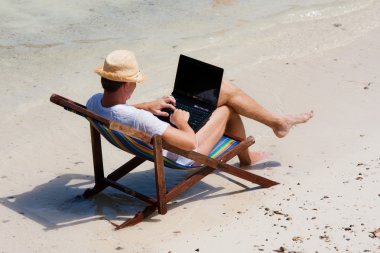 plajda şezlong ile bir dizüstü bilgisayar üzerinde oturan adam