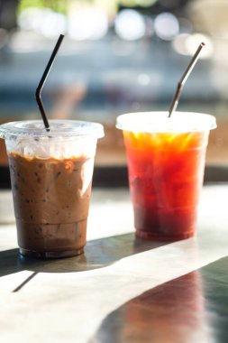 Plastik bir fincanda buzlu kahve (americano ve mocha kahve) ve ahşap masa üzerinde bulanık arkaplanlı kahverengi saman. Tatil ve rahatlatıcı konsept.