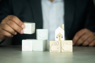 İşadamı ahşap bloğu şeffaf bir kumbara ikonu, cüzdan ikonu ve minyatür ahşap eviyle yönetiyor. Ev almak için para biriktirme, para biriktirmek için hedef plan