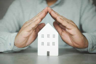 Wooden house covered by real estate insurance broker agent man hands protecting gesture of man and symbol of house. Property insurance and security concept.