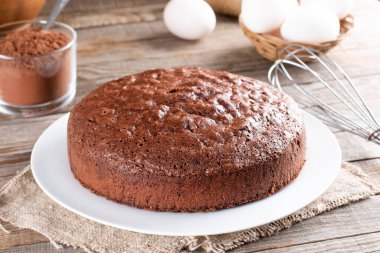 Homemade round chocolate sponge cake or chiffon cake on white plate so soft and delicious with ingredients: eggs, flour, cocoa, milk on wood table. Homemade bakery concept for background and wallpaper