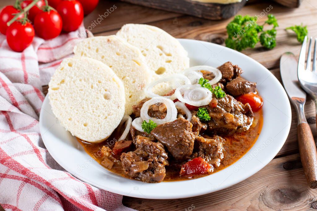 Pork goulash meat with dumplings on white plate, cutlery, garlic, onion, pepper, tablecloth in the background - typical Czech food