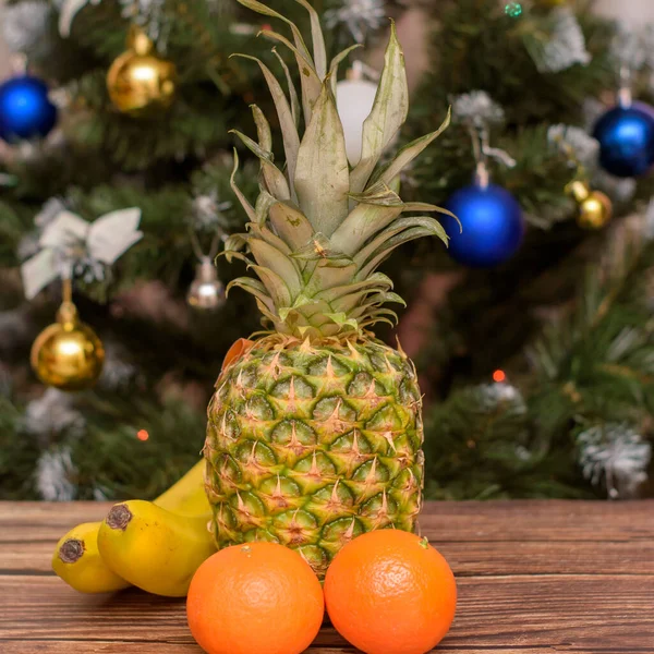 Año Nuevo y Navidad. Frutas tropicales sobre una mesa de madera. Mandarinas, plátanos y piña sobre un fondo de madera frente al árbol de Navidad. —  Fotos de Stock