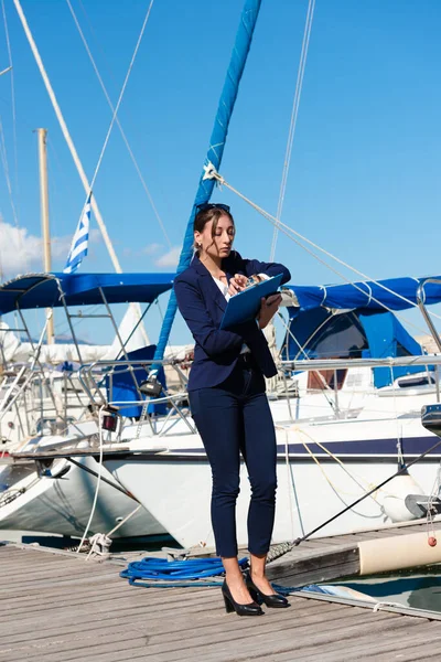 Distribuidor de yates hembra en traje azul marino sosteniendo bloc de notas, de pie fuera de un velero de lujo, mirando el reloj de pulsera. Heraklion, Creta, Grecia — Foto de Stock