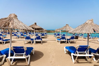 Sandalyeleri, güneşlikleri, saman şemsiyeleri ve mavi gökyüzü olan boş bir plaj. Akdeniz yaz tatili merkezi, Heraklion, Girit, Yunanistan.
