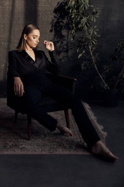 Siyah takım elbiseli güzel bir esmer. Aşağıya bakıyor. Gri arka plan ve saksı