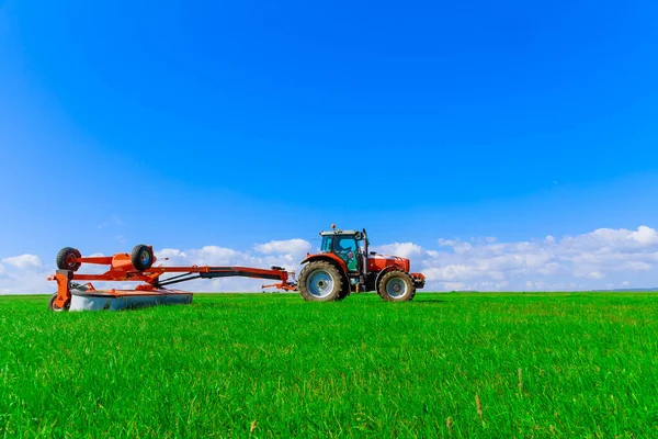 Traktor dengan mesin pemotong berputar memotong rumput di atas latar langit biru. — Stok Foto