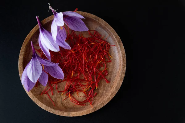 Three crocus flowers with saffron stamens on a wooden plate. Saffron stamens and flowers on a black background. — Fotografia de Stock