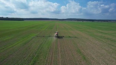 A tractor drives across a field, spraying fertilizer on a large field next to the road on which cars drive. Top view.