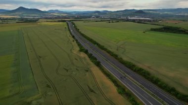 View from a height on the highway on which cars drive. Green fields near the road. Traces of tractor wheels in the fields.