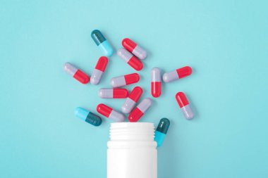 Mavi arka planda beyaz ilaç şişesinden sızan kapsül hapları.