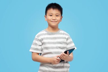 Elinde tabletle gülümseyen ve kameraya bakan mavi arka planda tişört giyen bir çocuk.