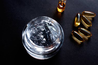 şeffaf kavanoz, nemlendirici krem, hiyalüronik asit ve kapsüllerdeki vitaminler siyah arka planda.