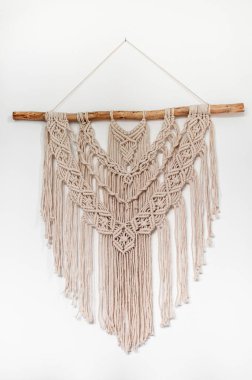 Beyaz arkaplanda bej rengi doğal ipliklerden el yapımı makrome örme tarzı bir duvar dekoru.