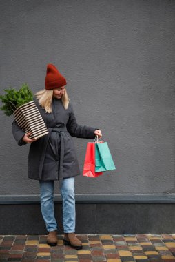 Kırmızı örgü şapkalı genç bir kadın Noel ve yeni yıl bayramları ve alışveriş torbaları için bir çuval köknar dalı taşıyor. Noel alışverişi