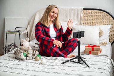 Genç ve güzel bir sarışın, Noel ve Yeni Yıl kutlamaları için ailesi ve arkadaşlarıyla görüntülü sohbet ediyor, geleneksel mavi ve kırmızı pijamalarıyla, rahat bir yatakta oturuyor, hediyeler ve tatil süslemeleriyle çevrili. Video çağrısı