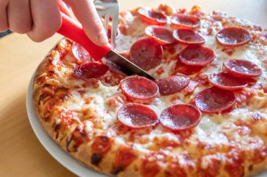 Oğlan elleri salam pizzasını çatal ve bıçakla parçalara ayırır pizza tabağında yakın çekim macro manzaralı lezzetli peynirli ve salamlı aç gençler için sağlıksız yağlı fast food olarak İtalyan leziz diyeti