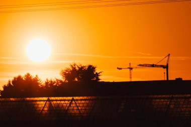 Turuncu gökyüzünde uzun vinç silueti, gökdelenler için yüksek voltaj hatları mimari takım çalışması olarak modern binalar ve şehir geliştirme mühendisliği ile inşaat alanı gösteriyor.