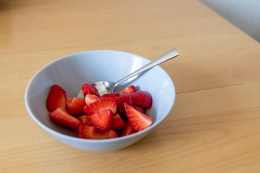 Kâsede kesilen çileklerin çoğu gümüş kaşıkla ya da lezzetli parmak yemekleriyle ve sağlıklı atıştırmalıklarla yenilebilir besin ve sağlık için ahşap mutfak masasında kırmızı meyve ve vitaminlerle yemeye hazırdır.