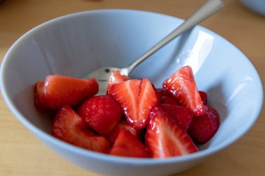 Kâsede kesilen çileklerin çoğu gümüş kaşıkla ya da lezzetli parmak yemekleriyle ve sağlıklı atıştırmalıklarla yenilebilir besin ve sağlık için ahşap mutfak masasında kırmızı meyve ve vitaminlerle yemeye hazırdır.