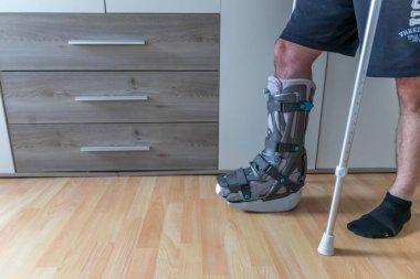 Achilles tendon yırtılması ameliyatından sonra Avrupalı bir adam evinde ay botu özel fizyoterapi ayakkabısıyla ve yaralı bacağa karşı evde tedavi için koltuk değnekleriyle ilk adımları PWB olarak atmayı öğreniyor.