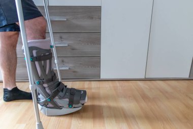 Achilles tendon yırtılması ameliyatından sonra Avrupalı bir adam evinde ay botu özel fizyoterapi ayakkabısıyla ve yaralı bacağa karşı evde tedavi için koltuk değnekleriyle ilk adımları PWB olarak atmayı öğreniyor.